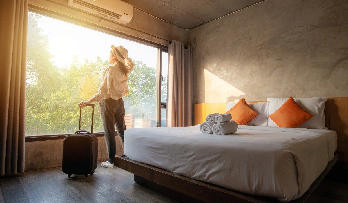 A traveler with a hat and suitcase beside a large window in a hotel room